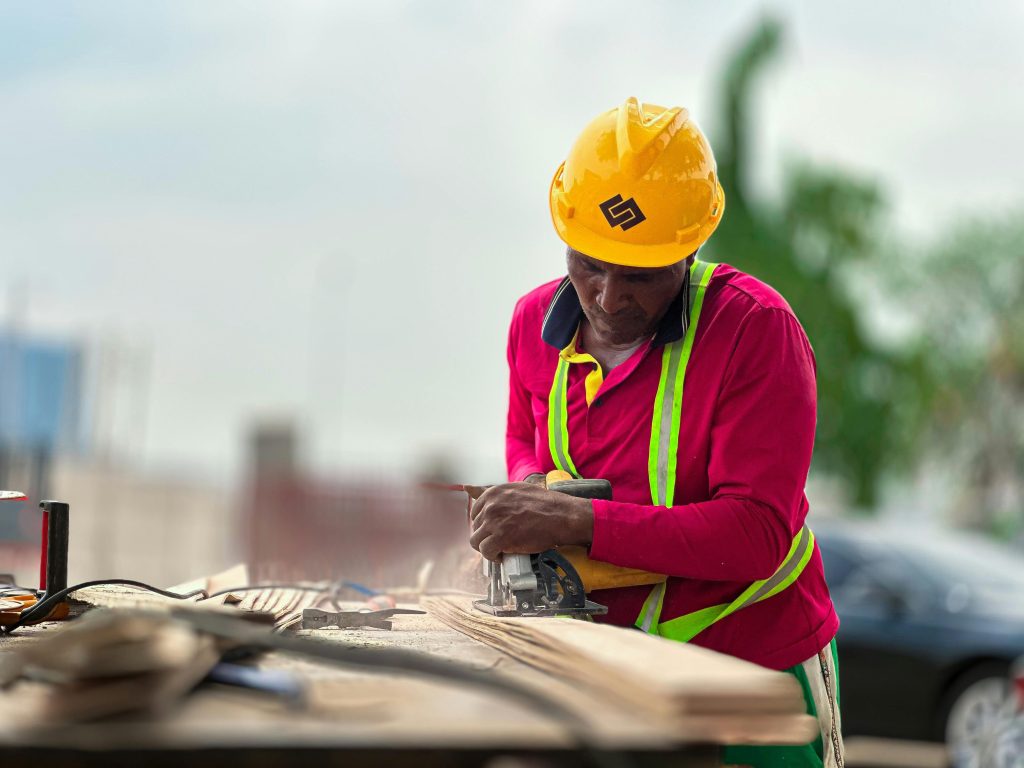 Construction worker cutting wood with a power tool outdoors, wearing safety gear.