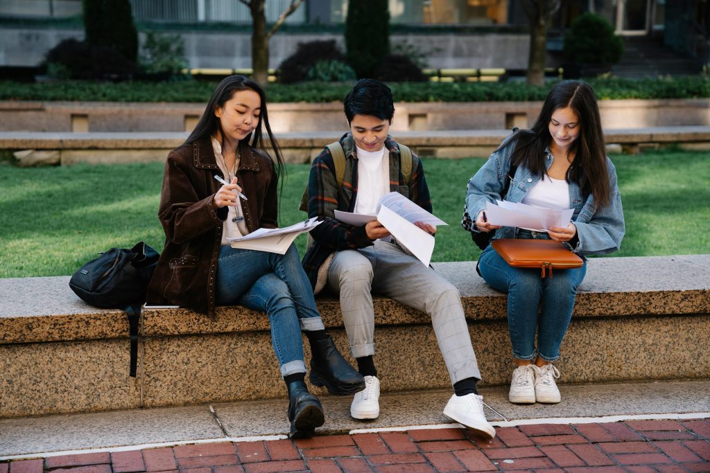 Three students studying together on a bench in a university campus setting outdoors.